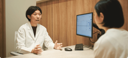 A doctor in a white coat speaking gently with a patient across a wooden desk